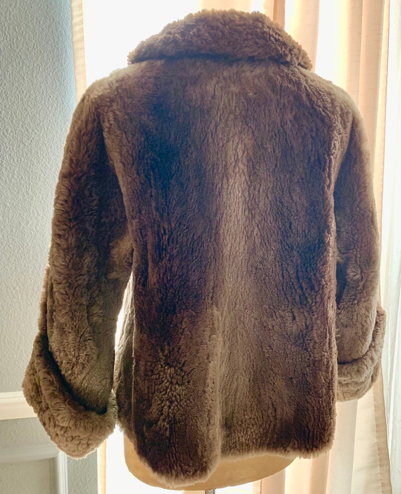 Pu&ograve; includere: Un cappotto marrone in finta pelliccia con ampio colletto e maniche lunghe. Il cappotto ha un aspetto morbido e strutturato e un design classico. Le maniche hanno un dettaglio con polsino arrotolato. Il cappotto &egrave; esposto su un manichino.