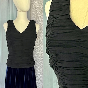 May include: A black sleeveless top with a V-neck and a ruched design. The top is displayed on a mannequin, paired with a dark blue velvet skirt. The fabric appears to be a soft, flowing material.