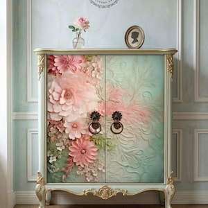 May include: A light blue cabinet with gold trim and ornate legs, featuring a large pink floral design on the left door and a textured, embossed leaf pattern on the right door. Two dark metal ring pulls are centred on the double doors.
