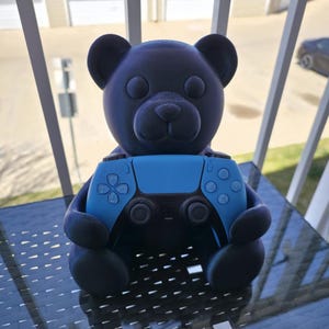 May include: A dark blue teddy bear figure holding a light blue video game controller. The bear has a textured surface and is sitting on a glass table. The controller has buttons and joysticks.