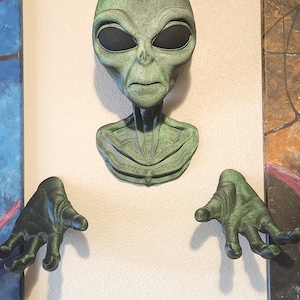 May include: A green alien head and two hands are mounted on a wall. The alien head has large black eyes and a textured surface. The hands are positioned on either side of the head, creating a unique decorative piece.