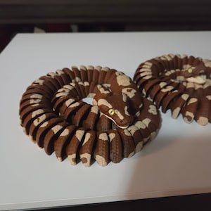 May include: Two articulated snake figurines featuring a segmented brown body with light beige patches. Each snake is coiled in a circular shape and has a detailed head with yellow eyes and a visible mouth. The surface has a textured scale pattern.