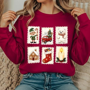 Może przedstawiać: Bordowa bluza z sześcioma świątecznymi wzorami w stylu znaczków pocztowych. Wzory obejmują elfa, czerwoną ciężarówkę z choinką, niedźwiedzia polarnego, piernikowy domek, skarpetę i świecę.