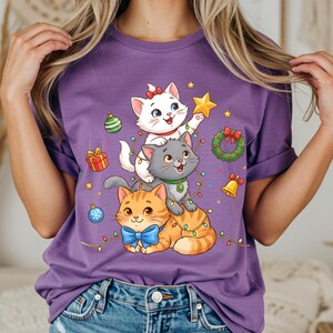 May include: Purple t-shirt featuring a festive design of three cartoon cats stacked on top of each other. The cats are decorated with Christmas ornaments, a star, and a wreath. The shirt has a rolled sleeve design.
