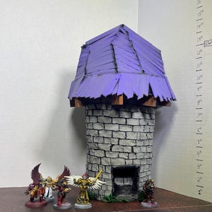 May include: A miniature fantasy tower with a purple roof and a stone brick facade. Several miniature figurines stand in front of the tower, including winged figures and armored warriors. The tower is a handmade model.