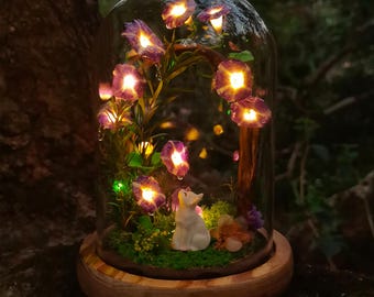 May include: A decorative glass dome featuring a miniature garden scene. Inside, illuminated purple flowers and a small white figurine are set against a backdrop of green moss and miniature plants. The dome rests on a wooden base.