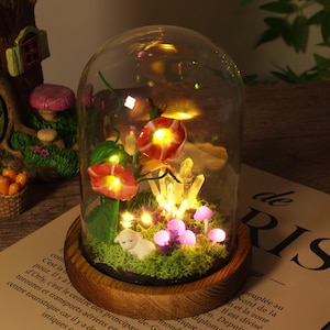 Könnte beinhalten: Dekorative Glaskuppel mit Holzsockel, mit beleuchteten roten Blumen, Kristallen und Miniatur-Lila-Pilzen. Eine kleine weiße Lammfigur ruht auf der moosigen Basis, wodurch eine skurrile Feengarten-Szene entsteht.
