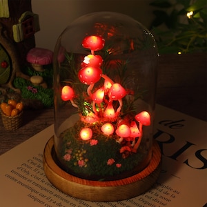 May include: A decorative glass dome featuring illuminated red mushroom figurines. The mushrooms are set on a bed of green moss and small flowers, with a wooden base. The dome is lit with warm, inviting lights.