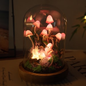 Puede incluir: Una cúpula de cristal decorativa con figuras de hongos rosas iluminadas. Los hongos están dispuestos entre vegetación y pequeñas luces, sobre una base de madera. La escena está iluminada con un brillo cálido, creando una estética caprichosa.