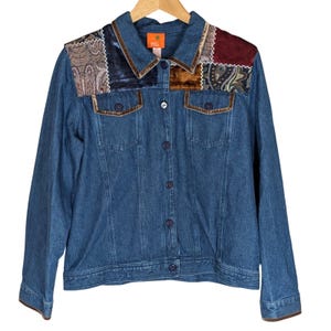 May include: A blue denim jacket featuring a button-down front, long sleeves, and two chest pockets. The upper shoulder area is accented with a patchwork design of velvet and paisley fabric in shades of burgundy, brown, and dark blue.