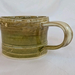 May include: A handmade ceramic mug with a rounded handle. The mug features a gradient of olive green and brown tones, with a white interior and handle. The mug has a rustic, textured appearance, suggesting it is hand-thrown.