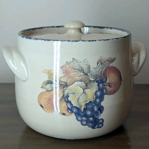 May include: Cream-colored ceramic cookie jar with a lid and two handles. The jar is decorated with a colorful illustration of apples, grapes, and leaves. The rim and lid have a blue trim. The jar is sitting on a wooden surface.