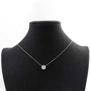 Può includere: Una delicata collana d'argento con un pendente rotondo e scintillante. Il pendente è centrato su una sottile catena, creando un design semplice ma elegante. La collana è esposta su un manichino nero.