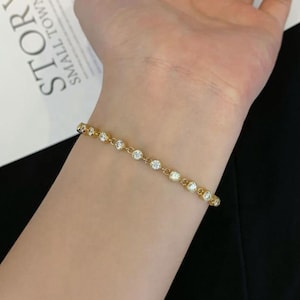 May include: A gold-colored bracelet with clear, round gemstones. The bracelet is delicate and sits on a wrist. The gemstones are evenly spaced along the length of the bracelet, creating a sparkling effect.