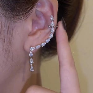May include: A silver ear cuff adorned with a cascade of shimmering, teardrop-shaped crystals. The cuff wraps around the ear, secured by a bar at the top. A row of crystals hangs down, adding to the elegant design.