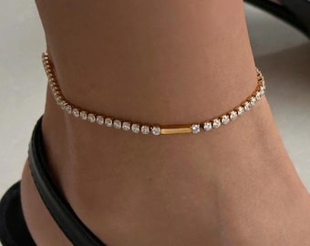 May include: A gold-toned anklet with a row of small, clear gemstones. The anklet is worn on an ankle, with a black sandal visible in the foreground. The gemstones are evenly spaced along the chain, creating a sparkling effect. The anklet's design is simple and elegant.