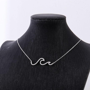 Puede incluir: Un collar plateado con un colgante en forma de ola. El colgante está adornado con pequeñas piedras brillantes, creando un efecto reluciente. El collar se exhibe en un maniquí negro, resaltando la delicada cadena y el colgante.