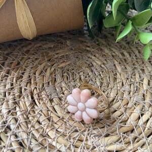May include: A gold ring with a pink flower-shaped stone. The ring is on a woven surface.