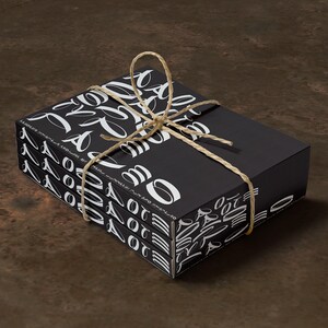 Puede incluir: Una pila de tres cajas negras con letras blancas, atadas con un cordel. Las cajas están etiquetadas con "O" y "OT" y presentan un patrón de letras estilizadas.