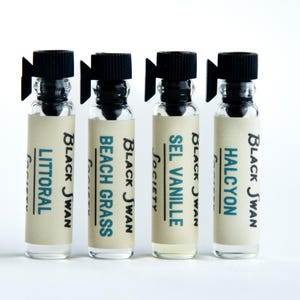 May include: Four small glass vials with black caps and labels. The labels read "LITTORAL", "BEACH GRASS", "SEL VANILLE Society", and "HALCYON" in a mix of black and teal text. The vials are arranged in a row against a white background.