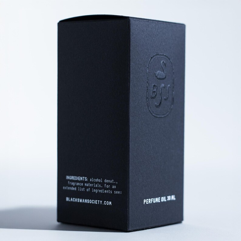 May include: A black perfume oil box with embossed logo and text. The box features the text "INGREDIENTS: alcohol denat., fragrance materials. For an extended list of ingredients see: BLACKSWANSOCIETY.COM PERFUME OIL 30 ML."