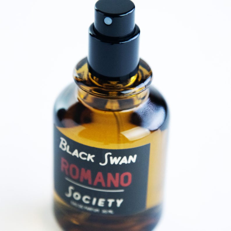 May include: A perfume bottle with a black cap and a brown glass body. The label reads "Black Swan Romano Society" in white and red text. The bottle is filled with a golden liquid.
