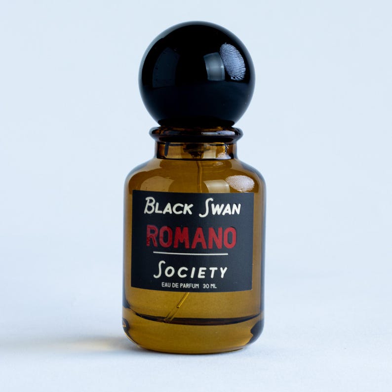 An apothecary style amber perfume spray bottle with a black gloss ball cap sits on a white surface against a white backdrop. The perfume bottle is labeled in black with the text, "black swan society, Romano, eau de parfum 30ml".