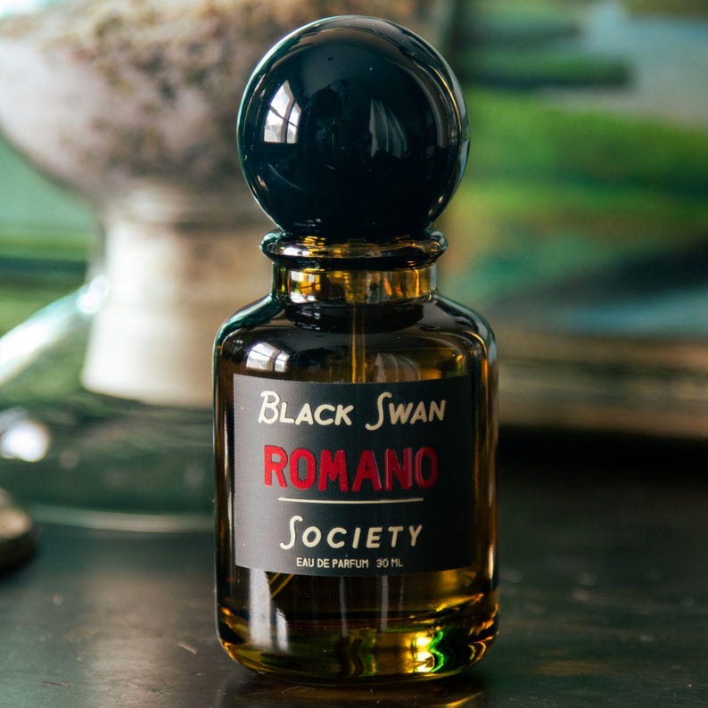 An amber perfume spray bottle with a black gloss ball cap sits on a dark wood surface lit by natural daylight. The background is out of focus but reveals a landscape painting and a glass specimen jar. The perfume bottle is labeled in black with the text, "black swan society, Romano, eau de parfum 30ml".