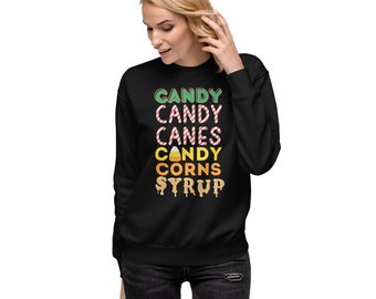 May include: A black long-sleeve sweatshirt featuring the text CANDY, CANDY CANES, CANDY CORNS, SYRUP printed in various colors. The word CANDY is green, CANDY CANES is red and white striped, CANDY CORNS is yellow and orange, and SYRUP is tan.