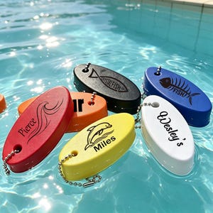 May include: A collection of oval-shaped foam floating keychains in red, orange, black, yellow, blue, and white. Each keychain features a metal ball chain and black printed designs including the names Pierce, Miles, and Wesley's, plus fish and dolphin graphics.