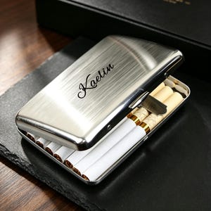 May include: A rectangular brushed silver metal cigarette case with a hinged lid. The top of the case features the name Kaelin engraved in a black script font. The case is partially open, revealing several cigarettes with white paper and gold filters inside.