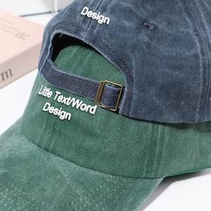 Puede incluir: Gorra de béisbol de dos tonos con la parte superior azul oscuro y la inferior verde. La palabra "Design" está bordada en la parte superior, y "Little Text/Word Design" está bordada en la parte posterior. La gorra tiene una hebilla de metal color latón.