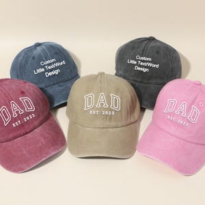 Op de afbeelding: Vijf baseballpetten in verschillende kleuren: bordeaux, blauw, grijs, beige en roze. De petten tonen het woord "DAD" in witte blokletters met "EST. 2023" eronder. Twee petten hebben de tekst "Custom Little Text/Word Design".