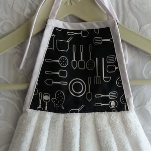 May include: White kitchen towel with black and white kitchen utensil print. The towel has a white ruffle and is hanging on a wooden hanger.