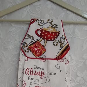 May include: White kitchen towel with a red border and a design featuring coffee cups, wine glasses, and the text "There's Always Time for Friends, Wine, and Coffee." The towel is hanging on a white hanger.
