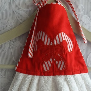 May include: A white terry cloth hand towel with a red top featuring candy cane and bow accents. The top is trimmed with red and white striped fabric and has a loop for hanging. The towel is on a wooden hanger.