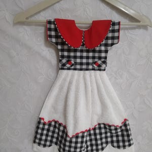 May include: A decorative kitchen towel shaped like a dress, hanging on a white hanger. The top features a red collar and black and white gingham pattern. The skirt is white with a gingham and red scalloped hem.