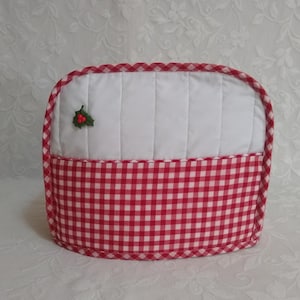 May include: A quilted tea cozy with a white top section and a red and white checkered bottom. A small holly sprig with red berries is attached to the white section. The cozy has a rounded top and is designed to keep tea warm.