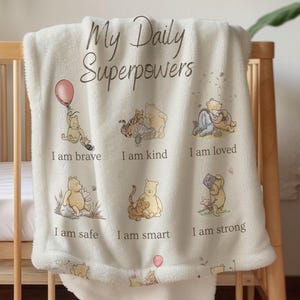 Puede incluir: Manta de bebé color crema con el texto "My Daily Superpowers" e ilustraciones de personajes de Winnie the Pooh. La manta presenta las frases "I am brave", "I am kind", "I am loved", "I am safe", "I am smart" y "I am strong".