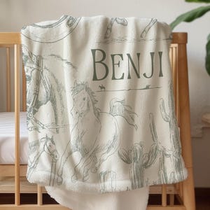 Può includere: Una coperta per bambini color crema con un tema western, con illustrazioni di cavalli, cactus e un cowboy. Il nome "BENJI" è stampato in verde. La coperta è drappeggiata su una culla di legno.