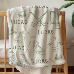 Puede incluir: Manta de bebé color crema con temática de hockey. La manta presenta dibujos lineales verdes de equipamiento de hockey, incluyendo patines, discos y palos, junto con el nombre "LUCAS" repetido.