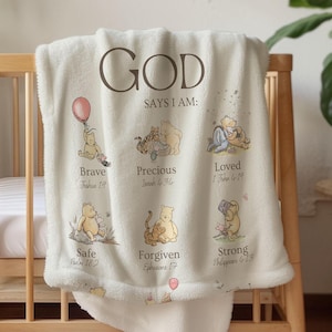 May include: Cream baby blanket with the text "GOD SAYS I AM" and illustrations of a bear. The blanket features the words "Brave", "Precious", "Loved", "Safe", "Forgiven", and "Strong". The blanket is draped over a wooden cot.