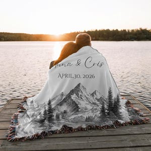 May include: A white blanket featuring a mountain and forest scene, personalised with the names "Anna & Cris" and the date "APRIL,30, 2026". The blanket has a fringed edge and is draped over two people on a wooden dock by a lake.