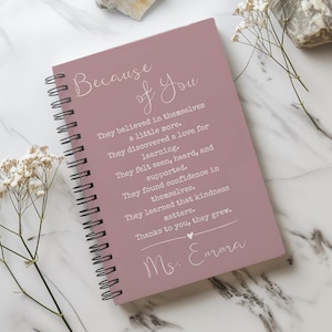 Puede incluir: Un cuaderno con espiral de color rosa polvoriento con el texto "Because of You". El cuaderno contiene un mensaje sobre el aprendizaje y la bondad. Flores blancas y una superficie de mármol completan la escena.
