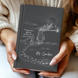 Puede incluir: Un diario de tapa dura gris oscuro con una ilustración de líneas blancas de Winnie the Pooh y Piglet caminando hacia unas montañas bajo un sol. El texto dice: "It makes such a difference," said Pooh, "To have someone who believes in you" y "Mr. Gordon."