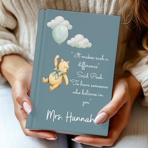 Puede incluir: Un libro personalizado con una cubierta azul que presenta una ilustración de Winnie the Pooh sosteniendo un globo. El texto dice "It makes such a difference" y "Mrs. Hannah". El libro tiene una cita sobre creer en alguien.