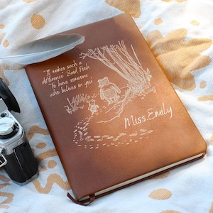 May include: A brown leather journal with the text "It makes such a difference Said Posh To have someone who believes in you Miss. Emily" and a Winnie the Pooh illustration. A white feather rests on the journal, next to a vintage camera.