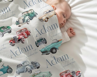 May include: A white baby blanket featuring watercolor illustrations of vintage cars in various colors. The blanket is personalized with the name "Adam" and the last name "Sheberton" in a blue script font. A baby is sleeping under the blanket.