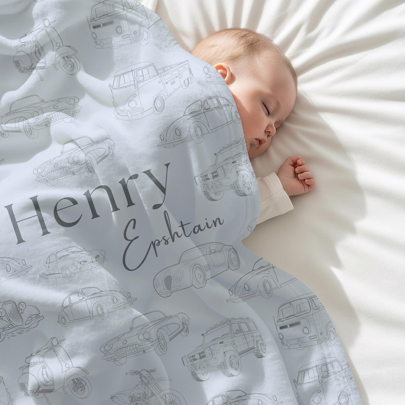 May include: A light blue baby blanket with a repeating pattern of vintage car illustrations in gray. The blanket features the name "Henry Epshtain" in gray script. A baby is sleeping under the blanket on a white surface.