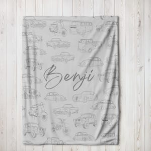 May include: A light gray blanket featuring a pattern of vintage cars, scooters, and vans in a line drawing style. The name "Benji" is written in a cursive font across the center of the blanket.